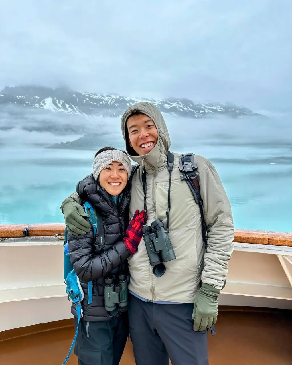 Deyu and Meesh bundled in winter jackets on a ship deck, a snow-streaked mountain and still glacial water behind them in Alaska.