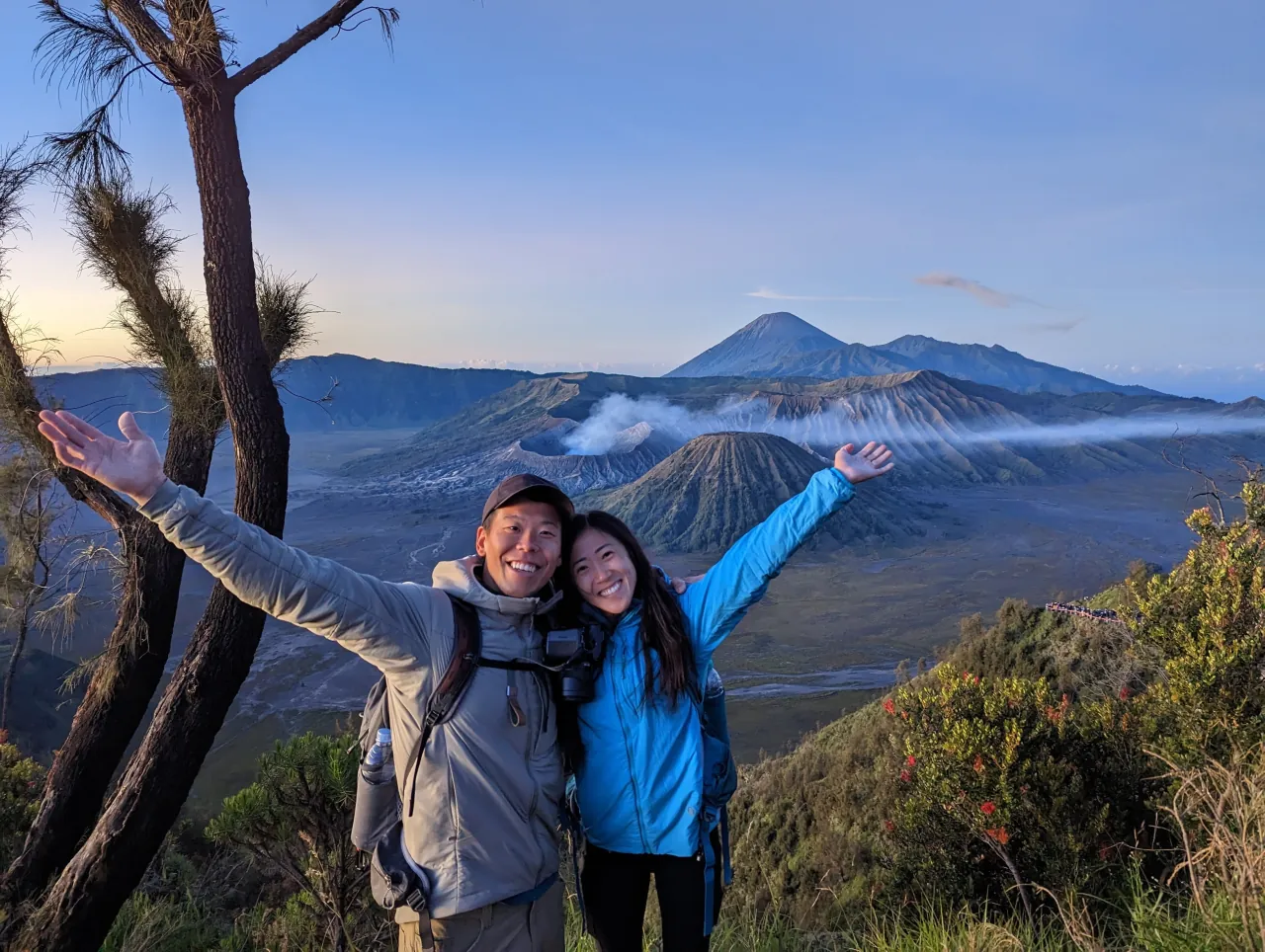 Deyu and Meesh with their arms raised on a ridge at sunrise, the smoking crater of Mount Bromo and distant volcanic peaks behind them in Java, Indonesia.