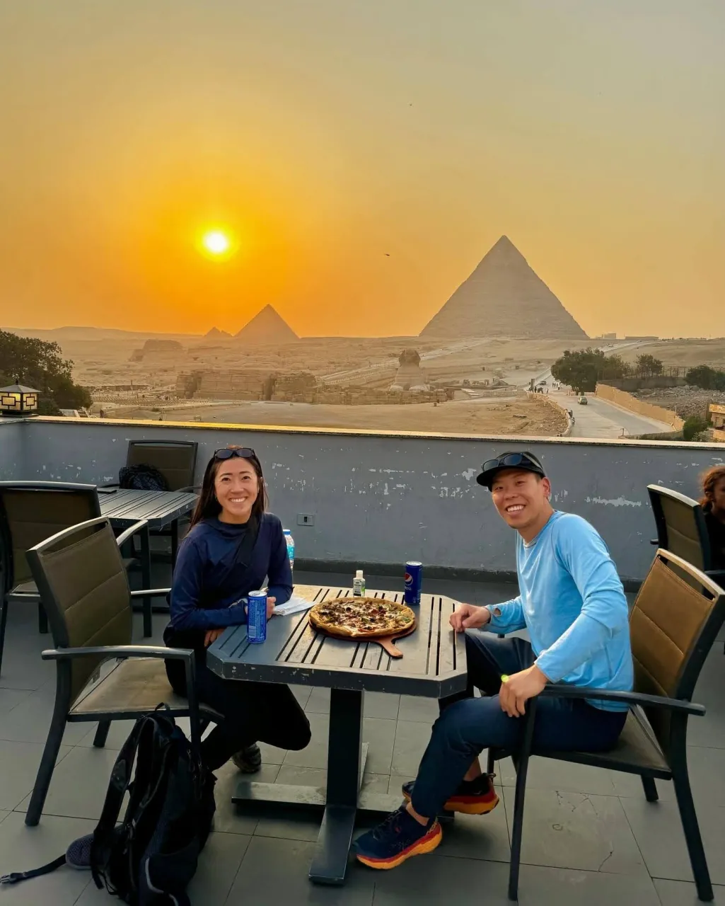 Deyu and Meesh at a rooftop table sharing a pizza as the sun sets behind the pyramids and Sphinx of Giza, Egypt.