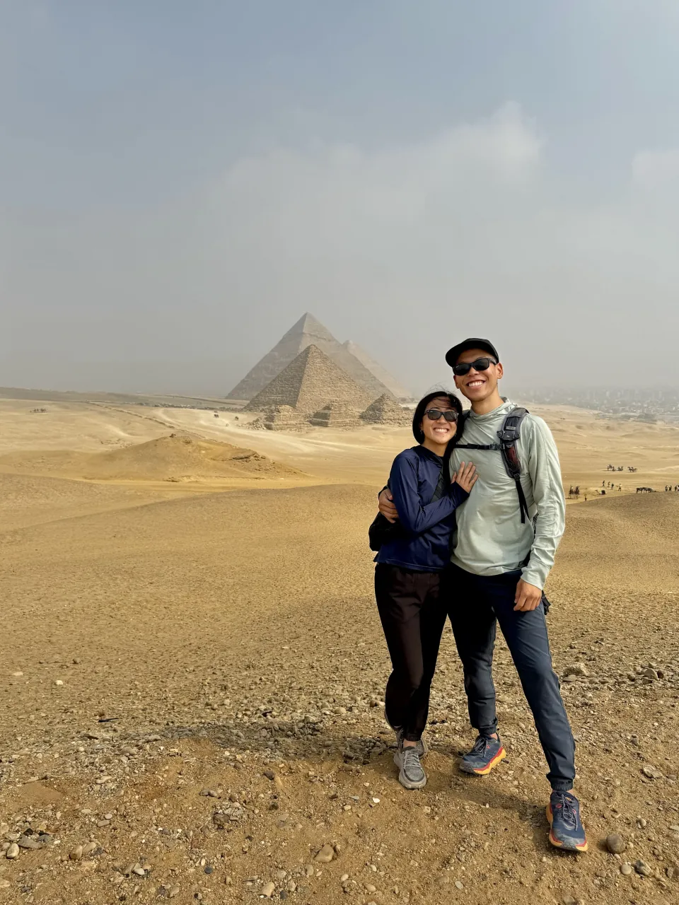 Deyu and Meesh standing in the desert with the three Pyramids of Giza rising behind them under a hazy sky in Egypt.