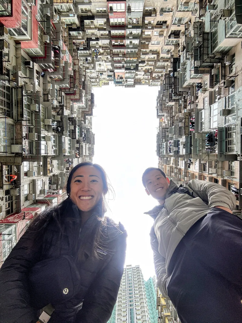 Deyu and Meesh looking up from the courtyard of a dense apartment complex, walls of stacked windows rising to a small square of sky in Hong Kong.