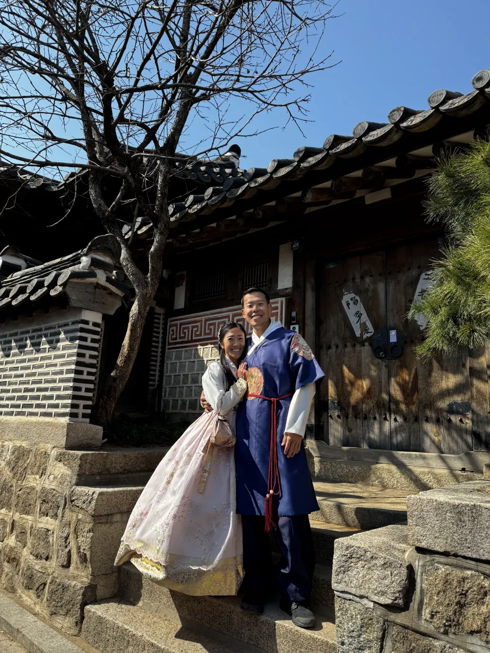 Deyu and Meesh wearing traditional hanbok in front of a wooden hanok gate on a sunny day in Seoul, South Korea.