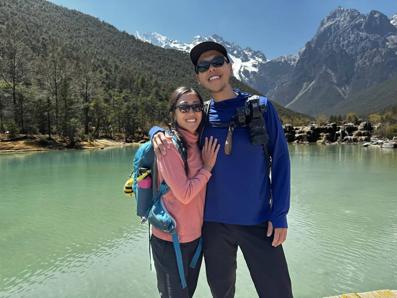 Deyu and Meesh at the edge of a bright turquoise pool with a snow-capped mountain rising behind the pines in Yunnan, China.